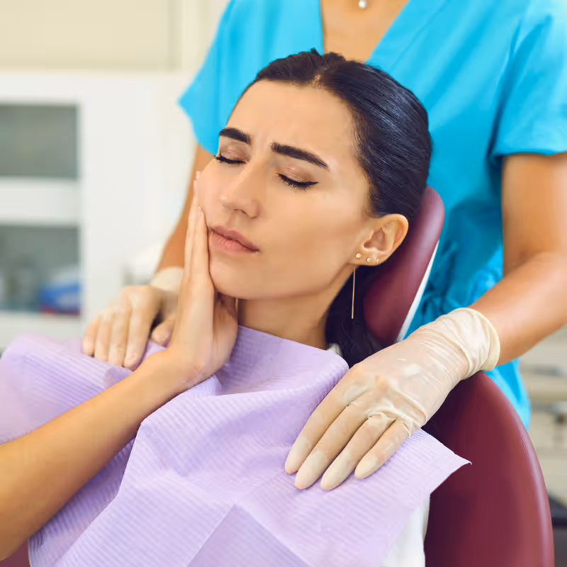 Woman sitting in dental chair holding her cheek in pain while a gloved dental professional comforts her.