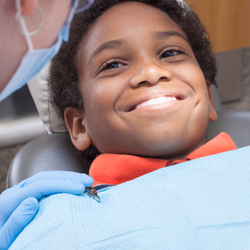 Smiling boy reclining in a dental chair with a dentist wearing blue gloves and a face mask attending to him.