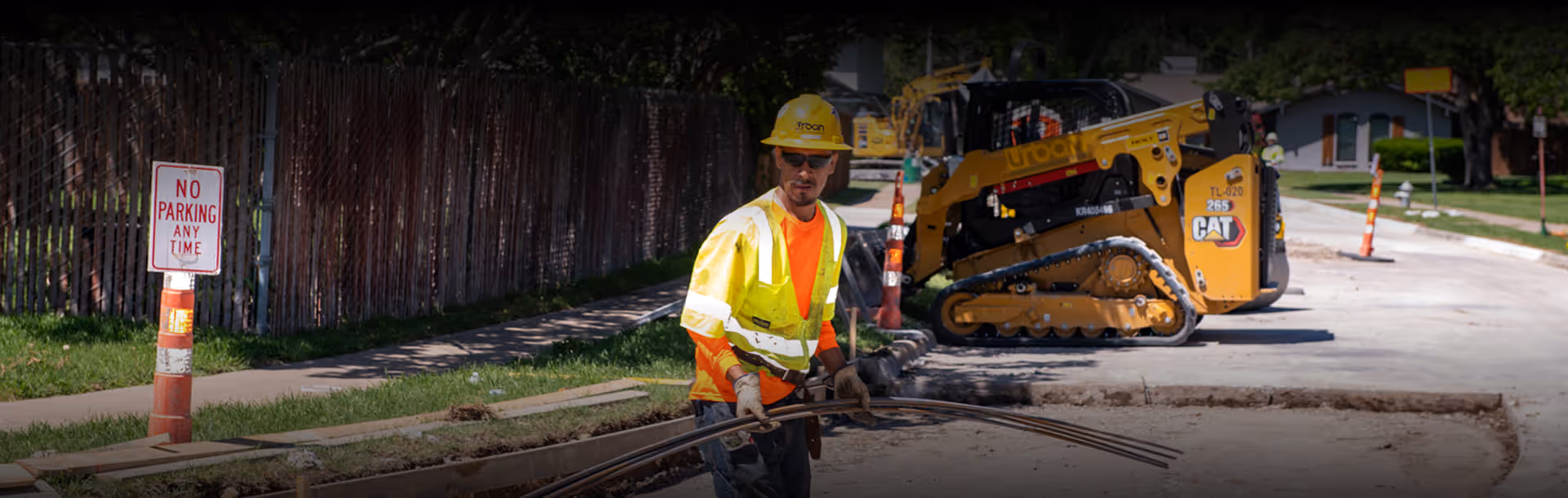 Construction worker wearing hard hat and safety vest carries metal rods on a road under repair with heavy machinery in the background.