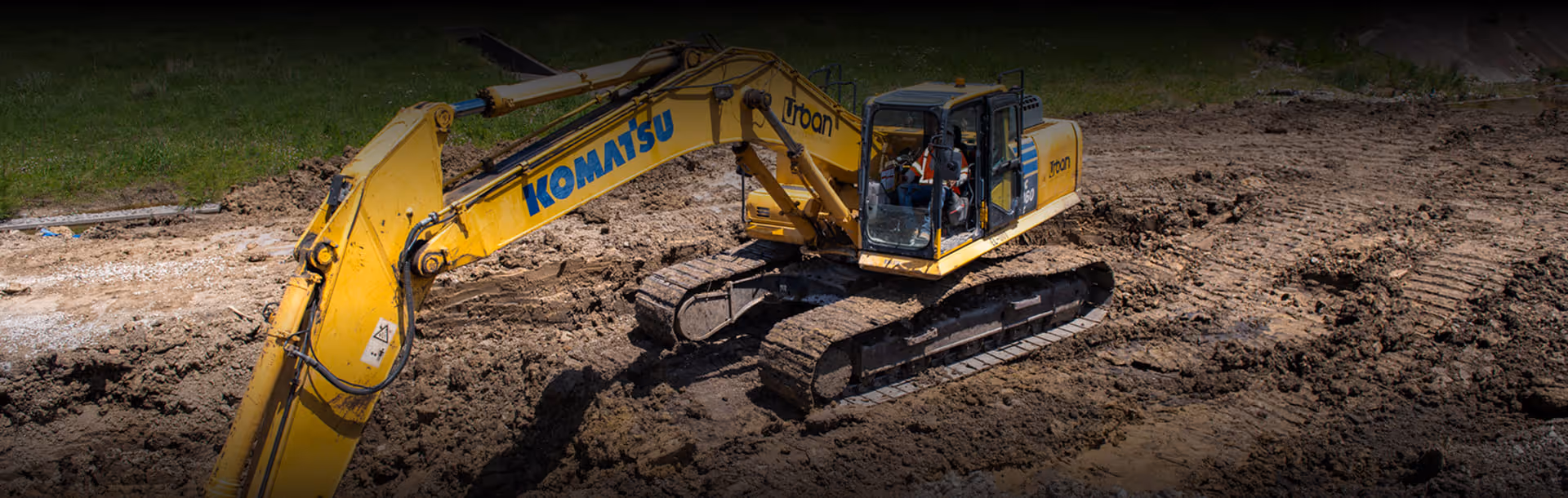 Yellow Komatsu excavator with Troan branding digging in a muddy construction site with grass in the background.