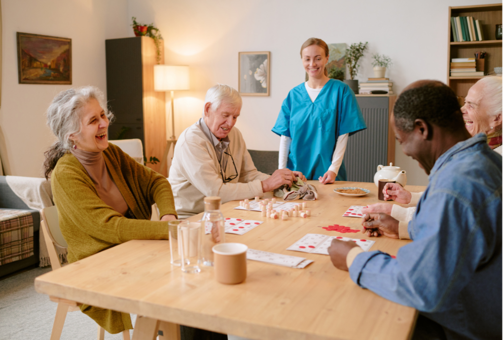 Group of elderly people sitting around a table playing bingo with a smiling caregiver standing nearby in a cozy, well-lit room.