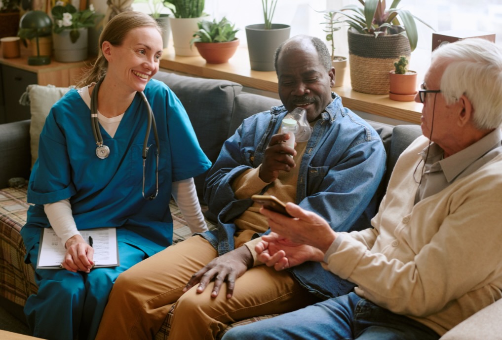 Healthcare professional chatting with two elderly men, one using an oxygen mask and the other holding a smartphone, seated on a couch.