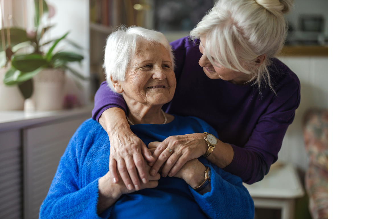 Elderly woman in blue sweater smiling while being embraced from behind by a woman with blonde hair wearing a purple sweater.
