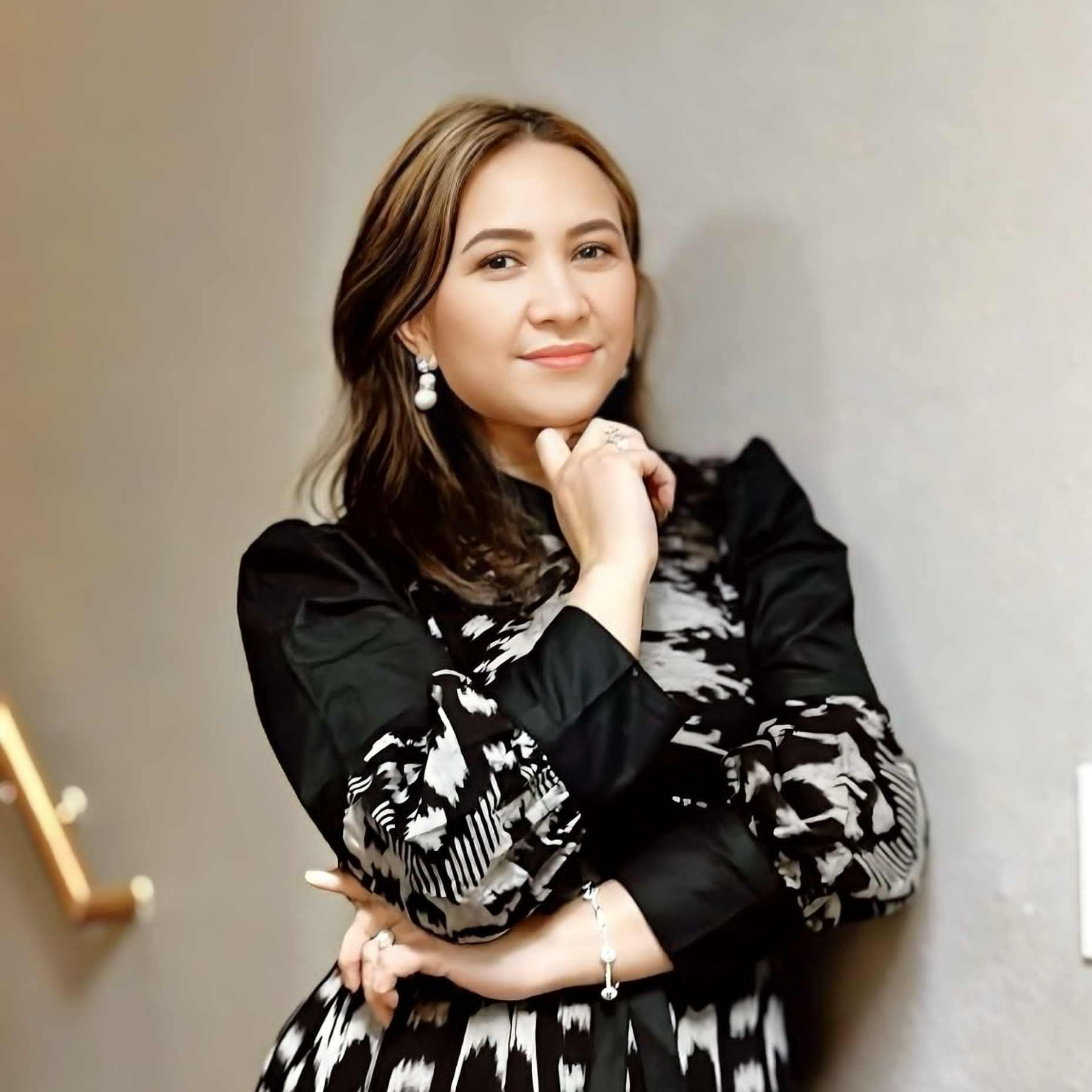 Woman in a black and white patterned dress posing with her arms crossed and hand resting on her chin, smiling softly.