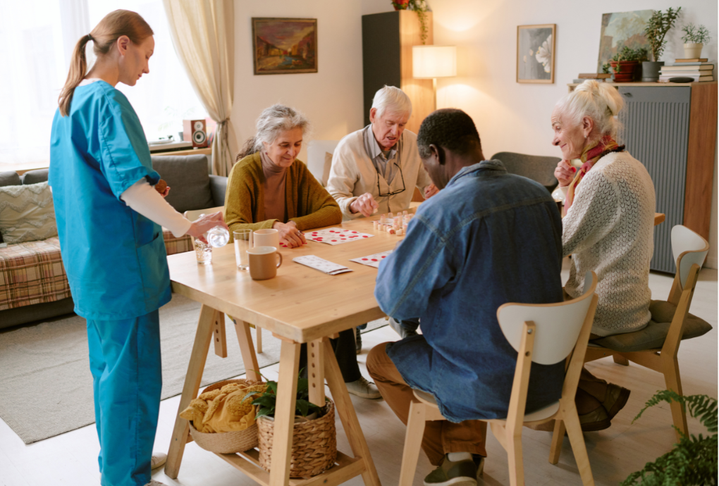 Caregiver pouring water for three elderly people playing a board game at a wooden table in a cozy living room.