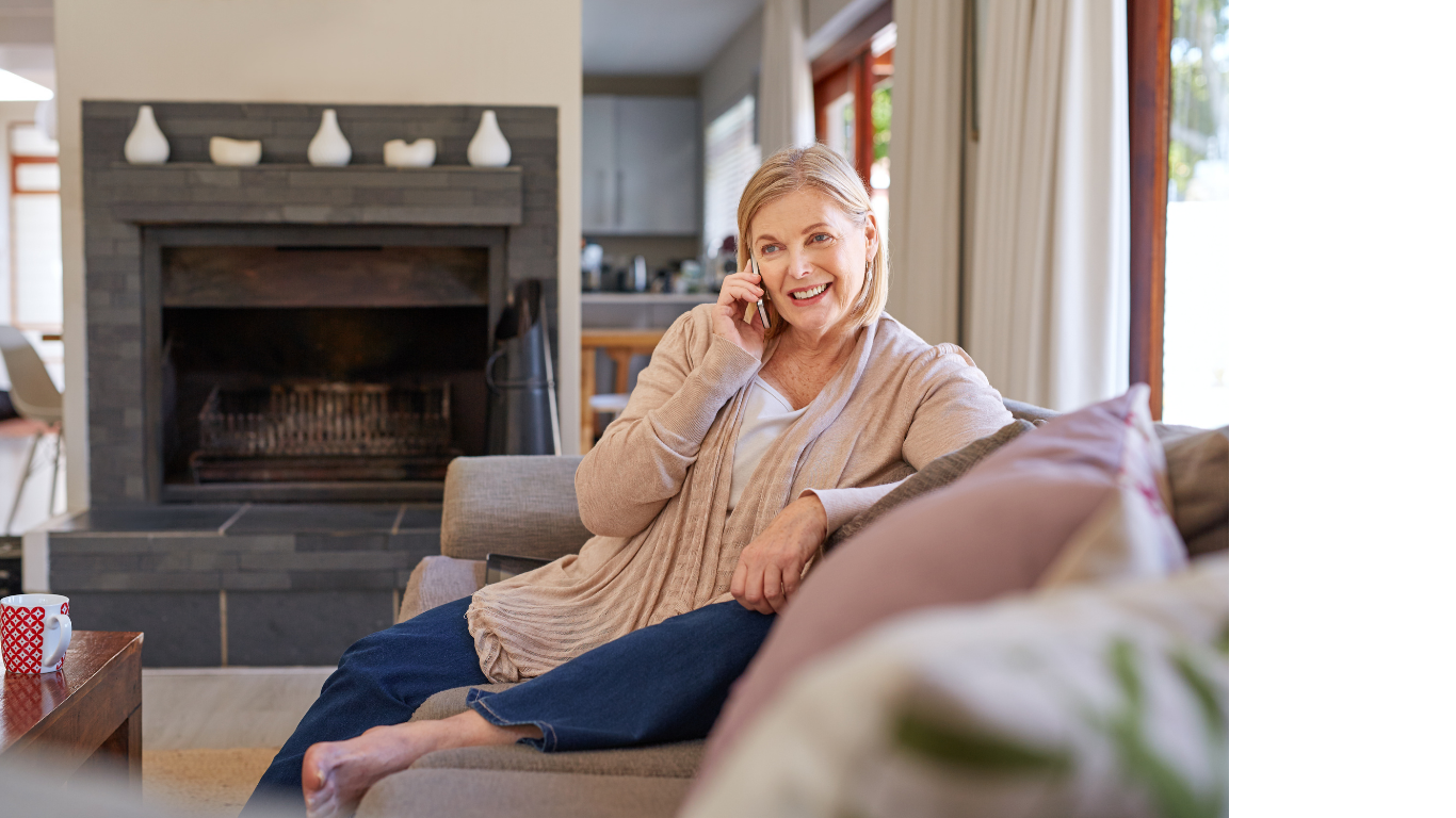 Smiling middle-aged woman sitting on a beige couch, talking on a smartphone in a cozy living room with a fireplace.