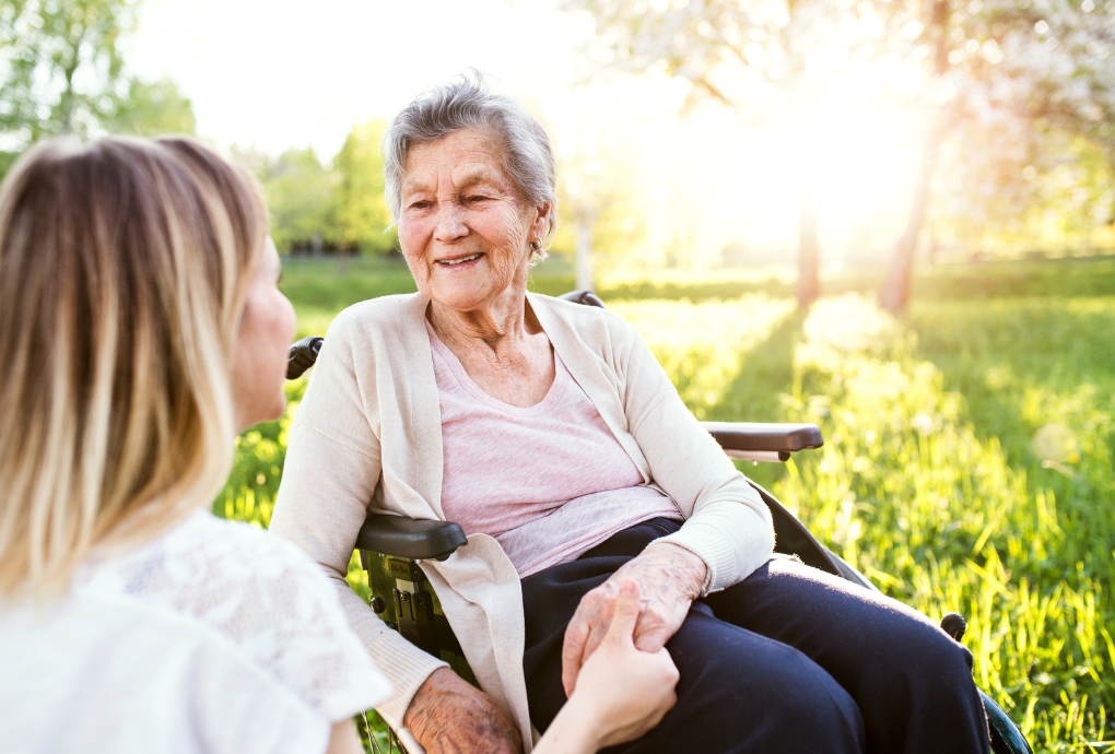 Older woman in a wheelchair holding hands and smiling with a younger woman in an outdoor sunny park.