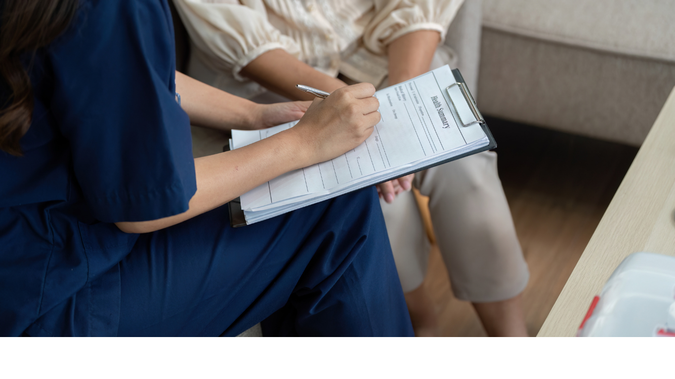 Healthcare professional in blue scrubs filling out a health summary form on a clipboard while consulting with a patient.