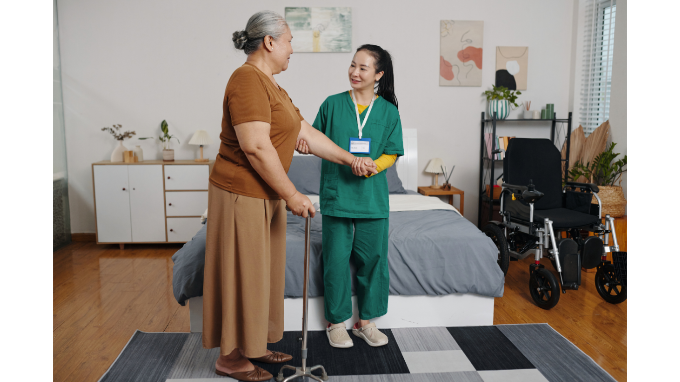 Caregiver assisting elderly woman using a walking cane in a bedroom with a wheelchair nearby.