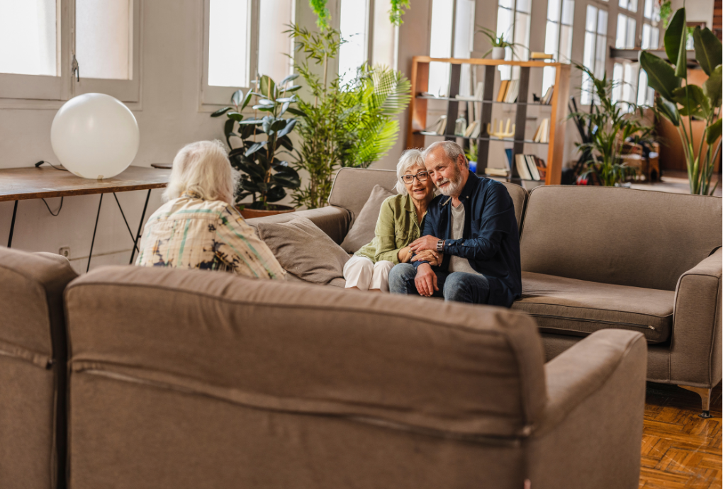 Two elderly people sitting on a sofa facing another elderly person in a cozy, plant-filled living room with large windows and bookshelves.