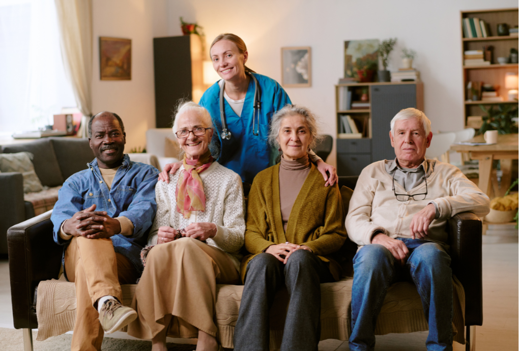 Smiling caregiver in blue scrubs stands behind a couch with four elderly people seated together in a cozy living room.