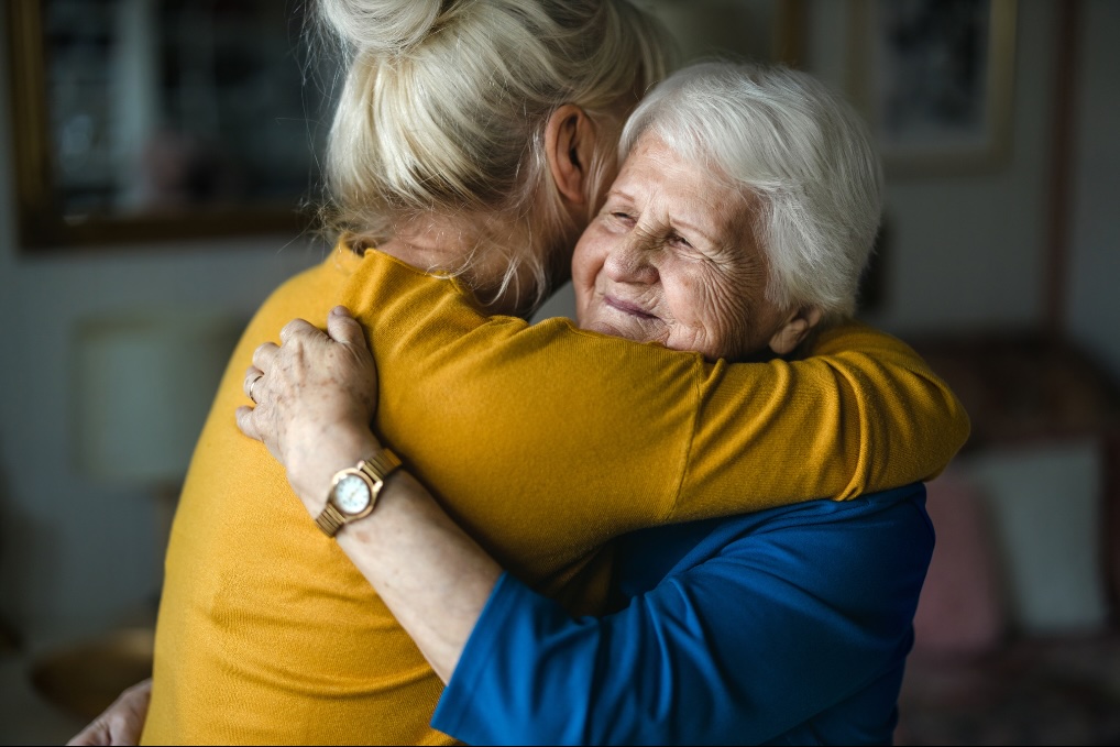 Two elderly women embracing warmly, one wearing a mustard yellow sweater and the other a blue top, sharing a tender moment indoors.