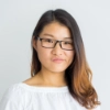 Young woman with long brown hair wearing black glasses and a white top against a plain light gray background.