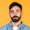 Portrait of a bearded man with dark hair wearing a denim shirt against an orange background.