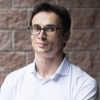 Man with short dark hair wearing glasses and a light blue collared shirt standing in front of a brick wall.