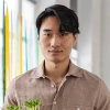 Portrait of a young man with dark hair wearing a beige shirt indoors, holding a small green plant.