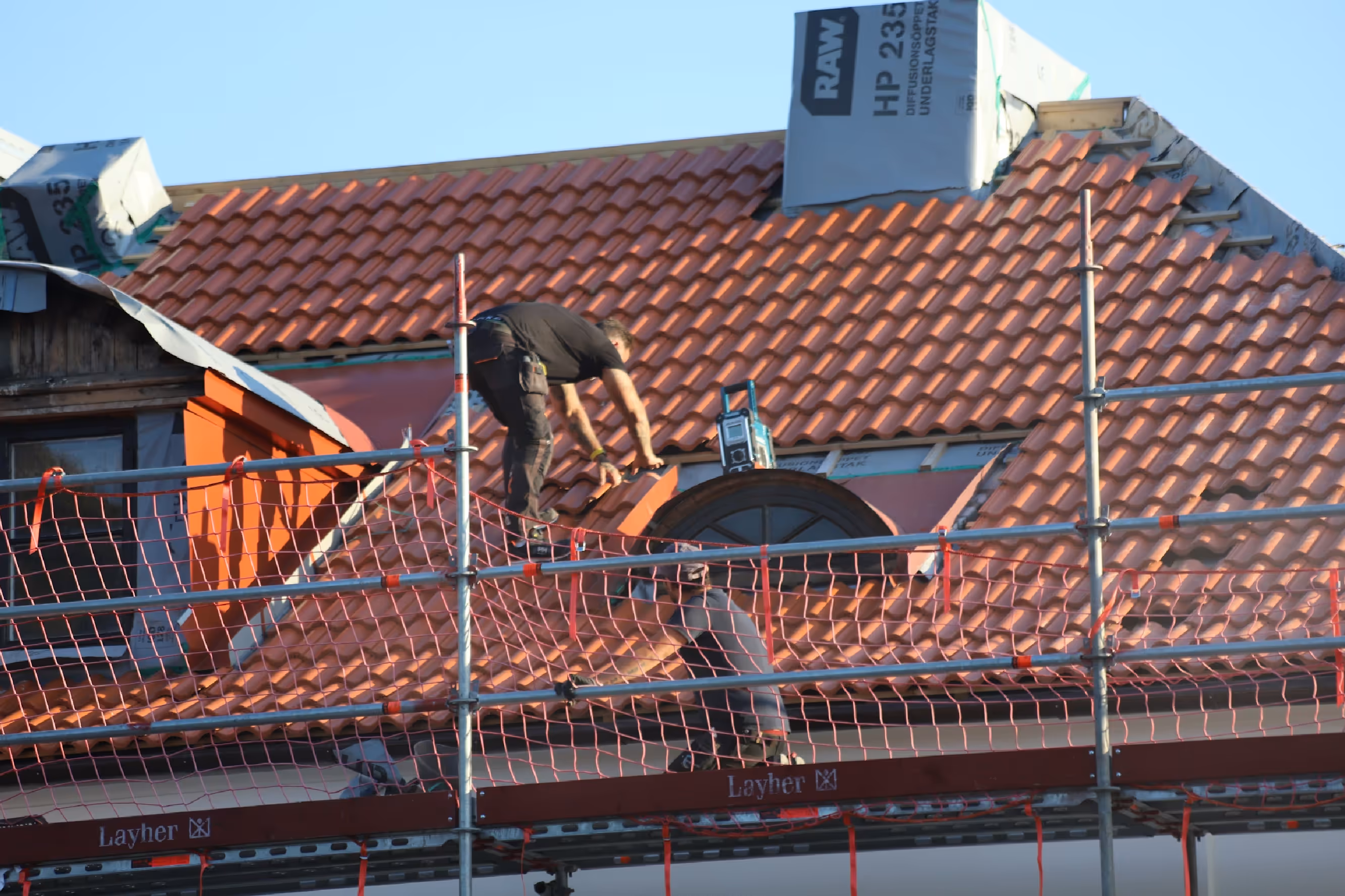 Two construction workers installing red roof tiles on a scaffolded house roof under clear sky.