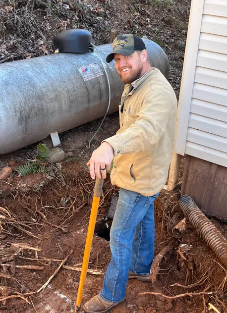 Jason McDaniel, owner/operator of Gold City Plumbing & Septic, holding a shovel by the corner of a house.