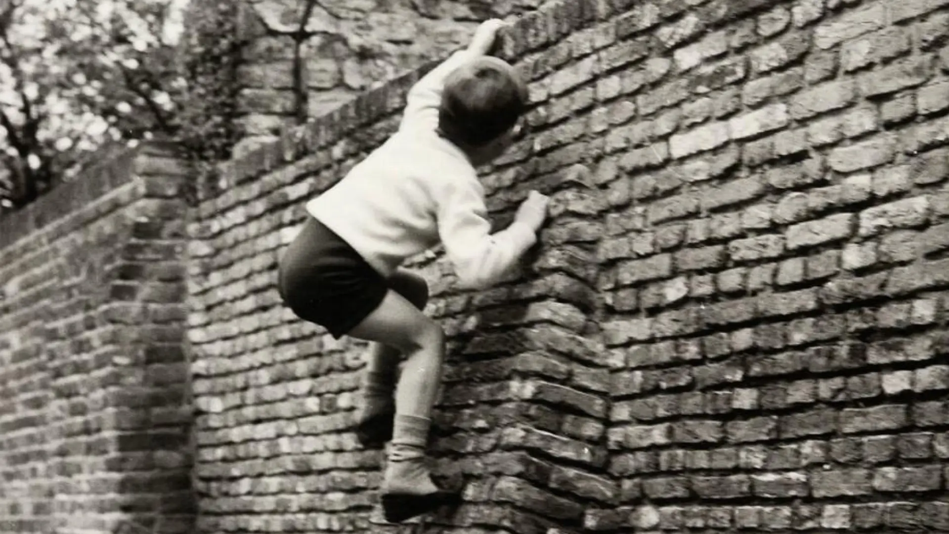 Child climbing a brick wall, captured in a black and white photograph.