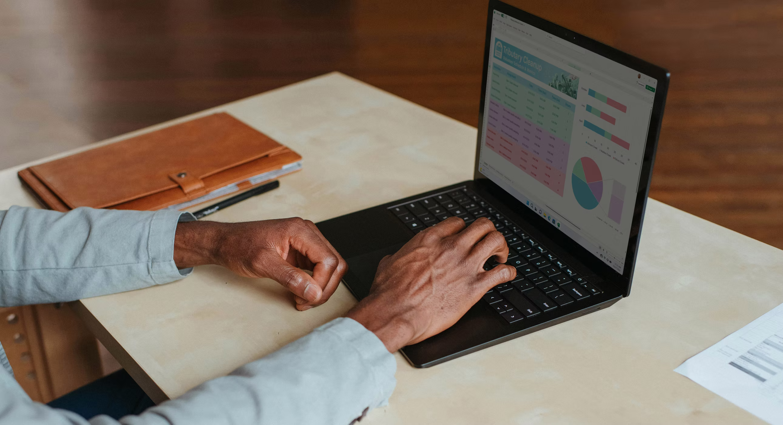 Person using a laptop displaying charts and data on a light desk with a brown folder and papers nearby.