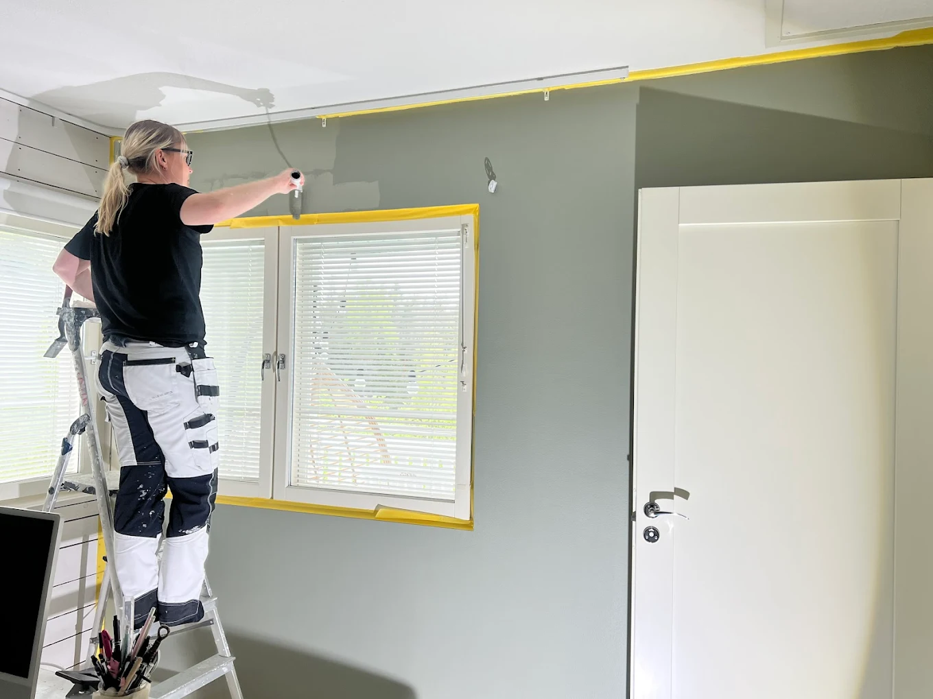 Nainen maalaa huonetta tikkailla, ikkuna ja ovi näkyvissä, valoisa huone.