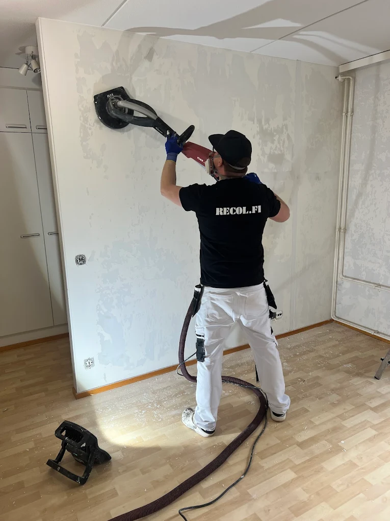 Mies hioo seinää hiomakoneella, jolla on letku pölynimuriin. Hänellä on yllään suojavarusteet, kuten hengityssuojain ja suojalasit. Lattialla on hiomapölyä.