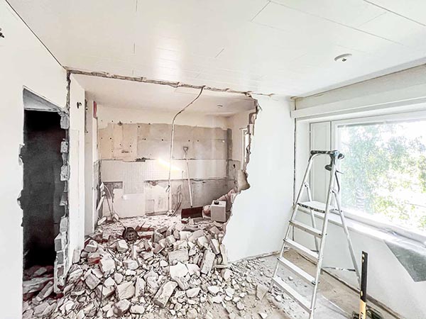 Kuva näyttää remontissa olevan huoneen, jossa on suuri kasa tiiliä lattialla, tikkaat ikkunan edessä ja seinät osittain purettu.