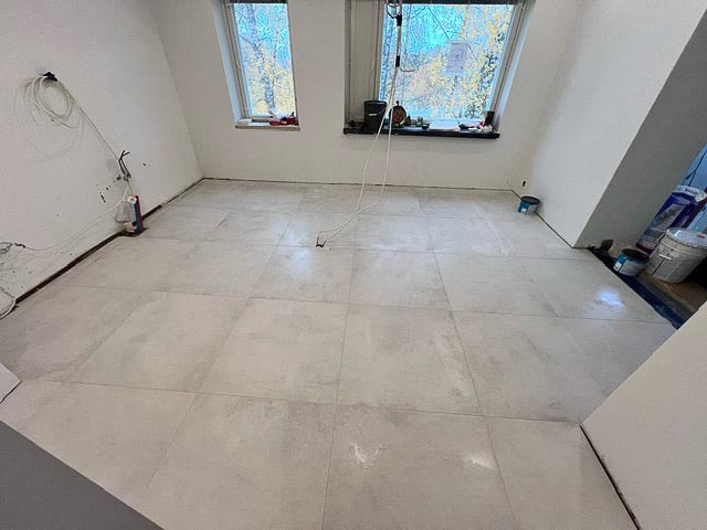 Huoneessa on vaaleat lattialaatat, kaksi ikkunaa ja valkoiset seinät. Sähköjohdot roikkuvat seinältä. Maalipurkkeja on nurkassa.