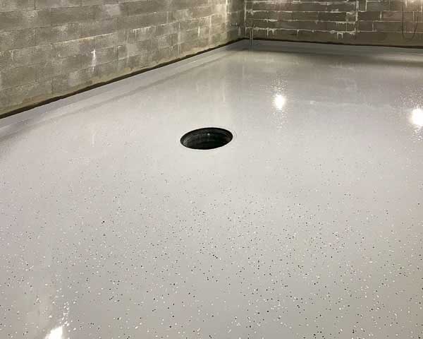 Vaaleanharmaa epoksilattia, jossa on pieniä mustia ja valkoisia pilkkuja, sekä pyöreä viemäri keskellä huonetta. Seinät ovat harmaita betoniharkkoja.