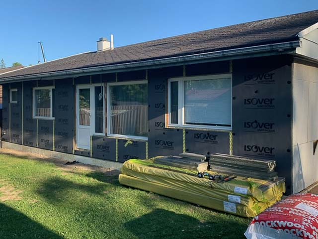 Yksikerroksinen talo, jossa on musta ulkoverhous, ikkunat ja ovi, sekä kasa rakennusmateriaaleja nurmikolla aurinkoisena päivänä.