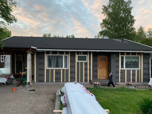 Yksikerroksinen talo, jossa on musta katto, harmaa ulkoverhous ja puinen etuovi. Lapsi seisoo etuoven edessä.