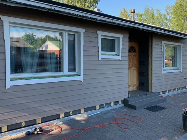 Vaaleanruskea puutalo, jossa on valkoiset ikkunakehykset ja tummanharmaat portaat, valoisa sää.