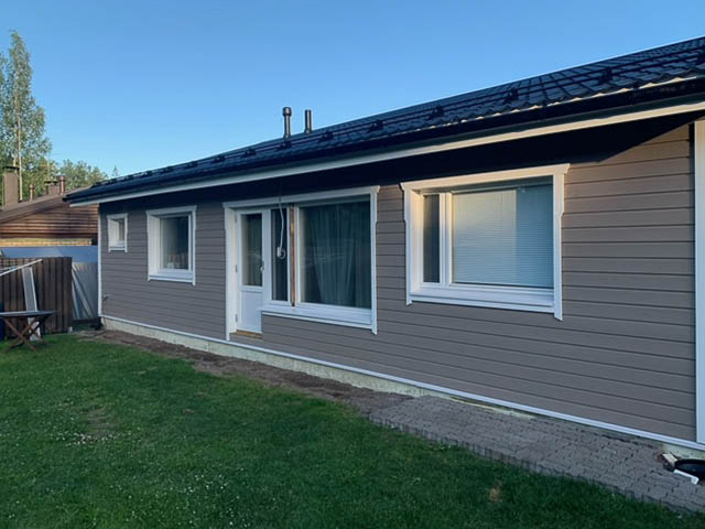 Yksikerroksinen harmaa talo, jossa on valkoiset ikkunakehykset, tumma katto ja vihreä nurmikko.