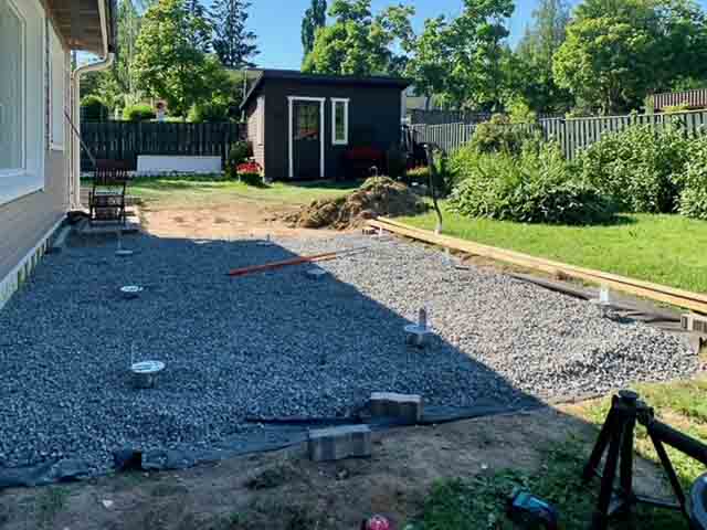 Ulkona on kivetty alue, jossa on rakennustyömaa, puutarha ja pieni rakennus taustalla. Aurinkoinen sää valaisee maisemaa.