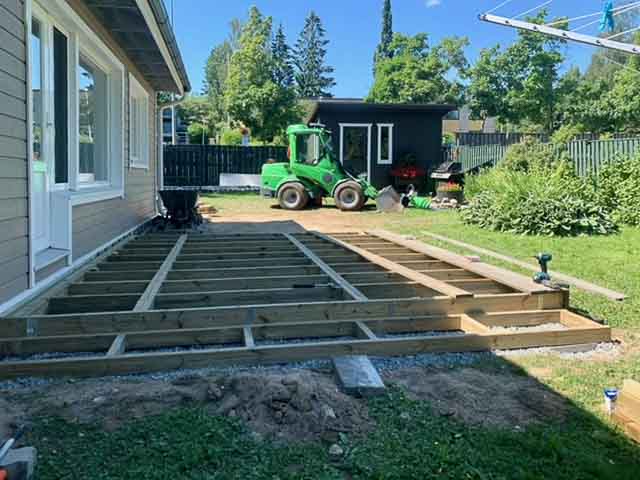 Ulkona on rakenteilla puinen terassi, jossa on näkyvissä puurunko ja portaat. Taustalla on vihreä traktori ja puutarhavaja.