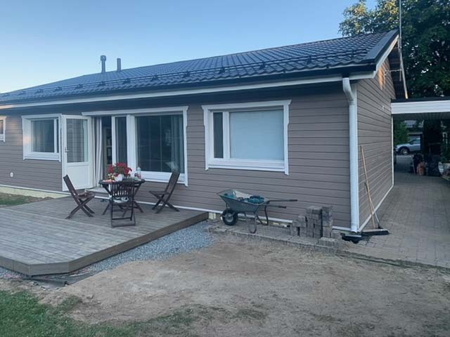 Ruskeasävyinen omakotitalo, jossa on harmaa katto, terassi ja puutarhakärry. Aurinkoinen sää valaisee maisemaa.