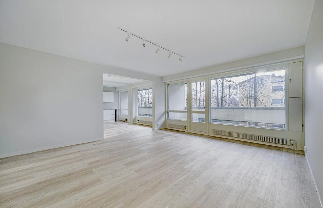 Vaalea, tyhjä huone, jossa on suuret ikkunat ja vaalea puulattia. Kattovalaisimet valaisevat tilaa.