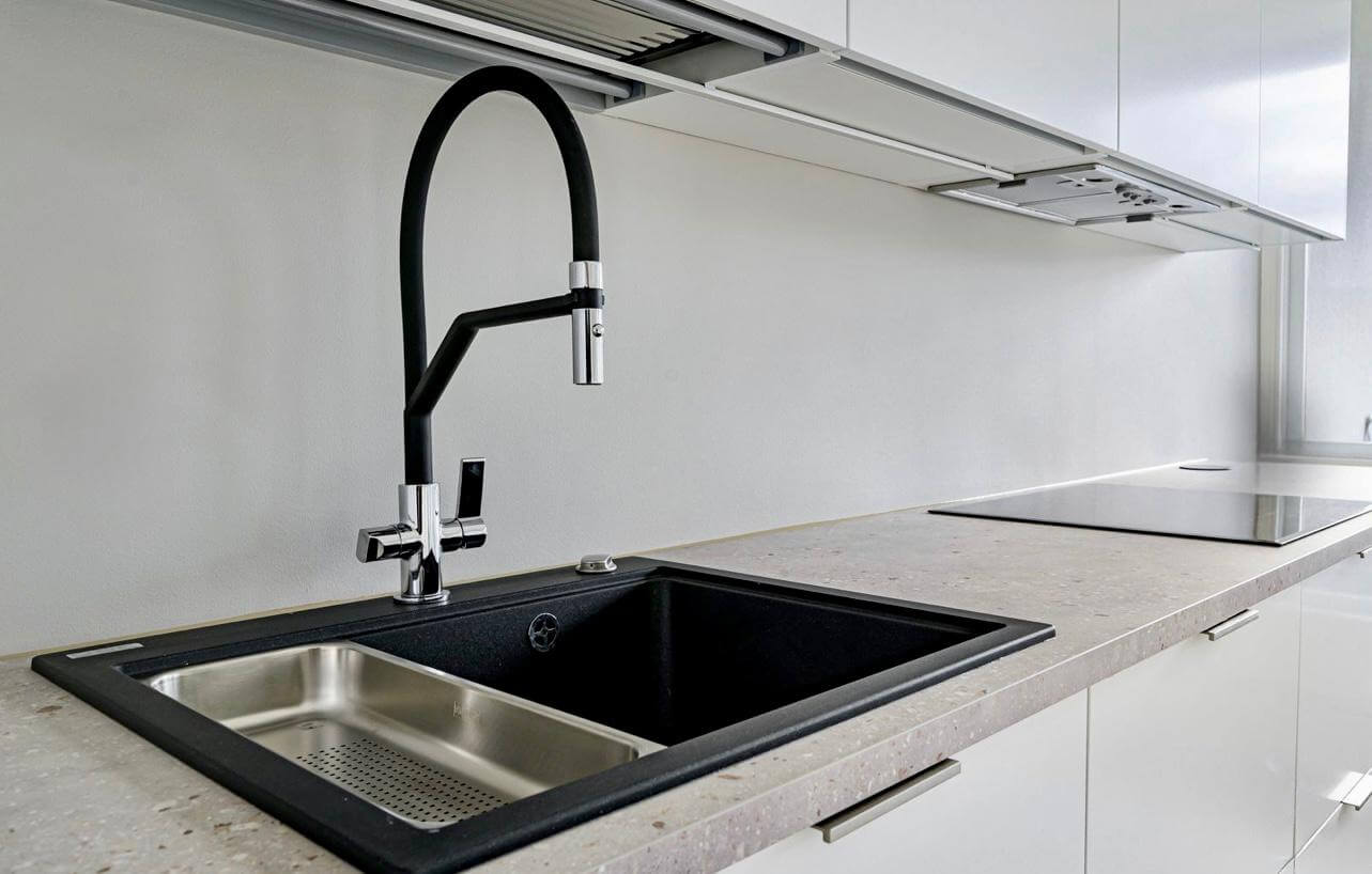 Keittiön musta pesuallas, kromihana ja harmaa työtaso valkoisessa keittiössä, jossa on moderni liesituuletin ja valoisa tunnelma.