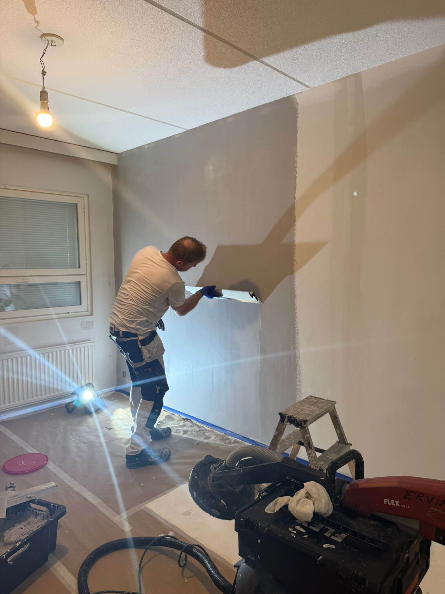 Sisätila, jossa mies tasoittaa seinää lastalla. Huoneessa on ikkuna, lattialamppu ja työkaluja. Seinä on osittain tasoitettu harmaalla massalla.