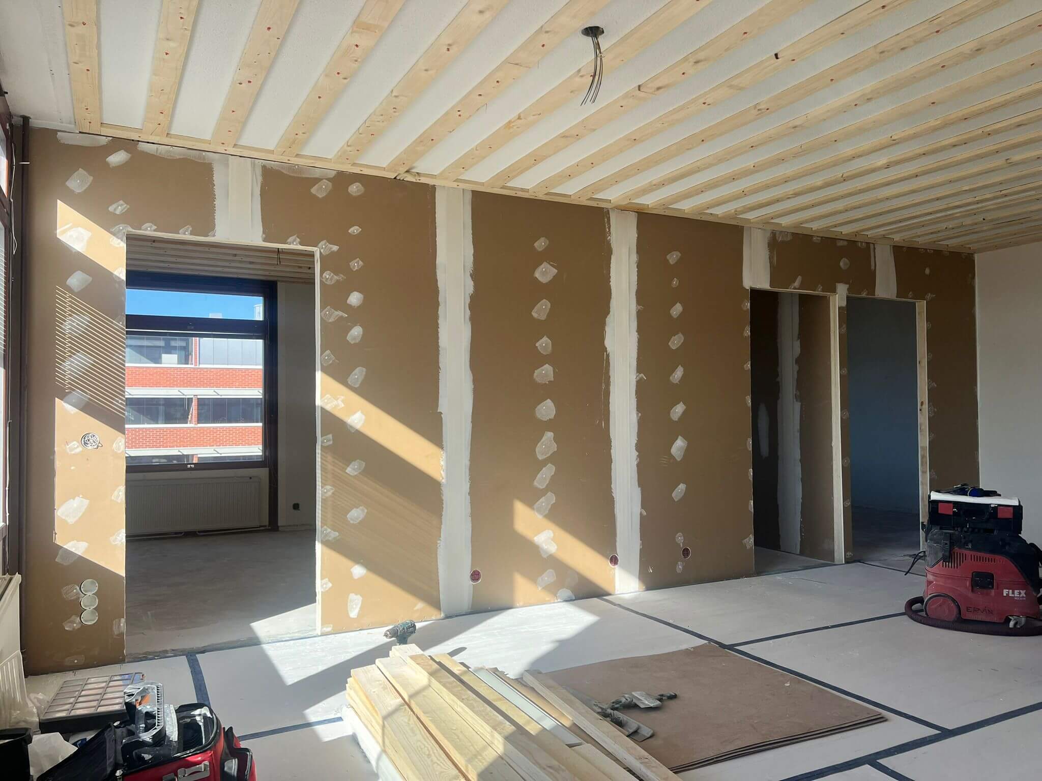 Rakennustyömaa, jossa on puupalkit katossa, kipsilevyseinät täytetyillä saumoilla, ikkuna ja työkaluja lattialla, valaistus on kirkas ja luonnollinen.