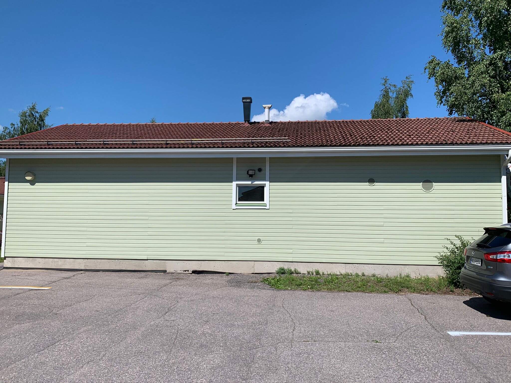 Vaaleanvihreä rakennus, jossa on ruskea katto, pieni ikkuna ja harmaa auto pysäköitynä asfalttipihalle aurinkoisena päivänä.