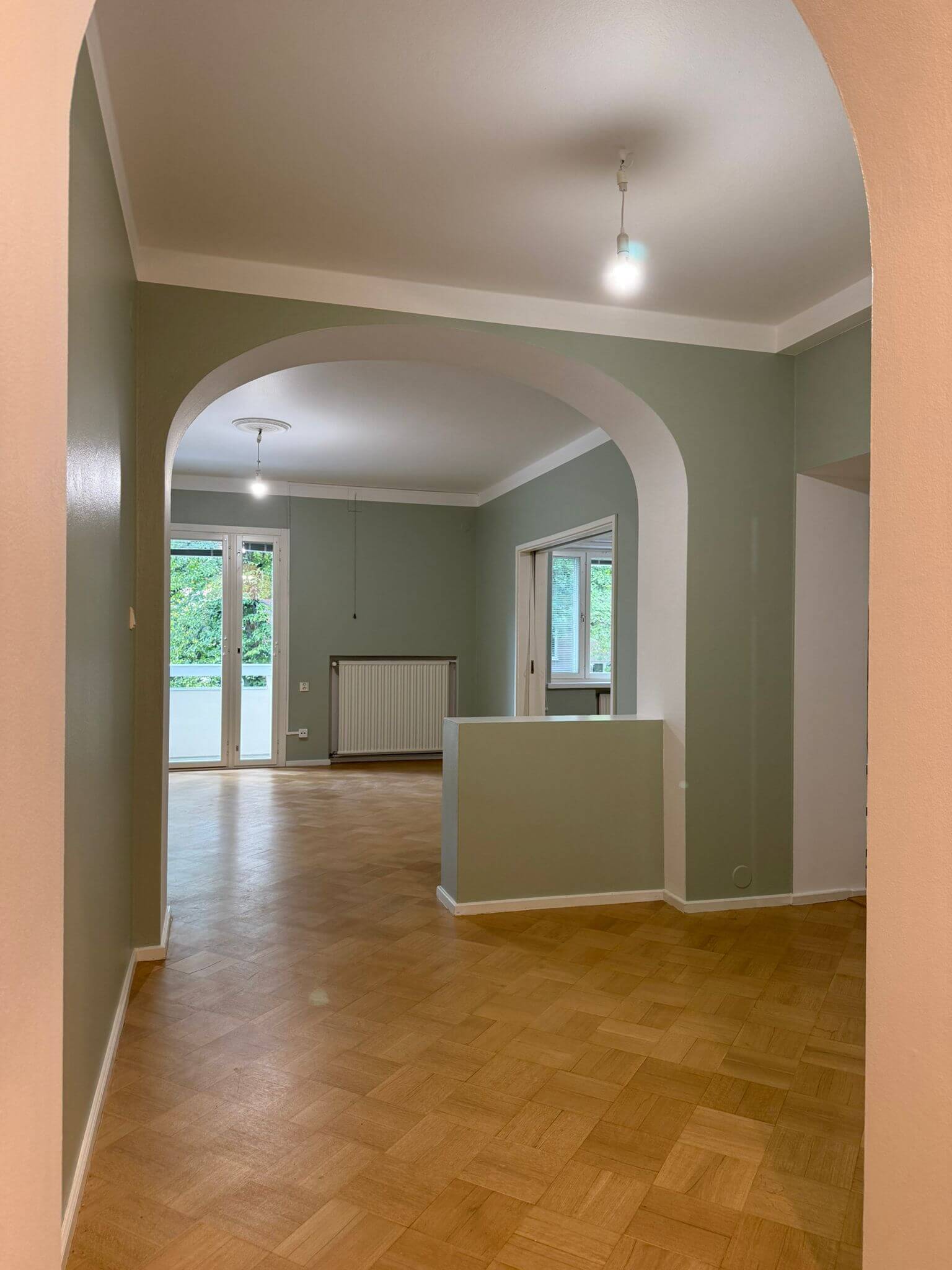 Huoneiston sisätila, jossa on parkettilattia, kaariholvit ja vihreät seinät. Huoneessa on valkoinen patteri ja ikkunat, joista näkyy ulos. Valoisa ja avara tila.