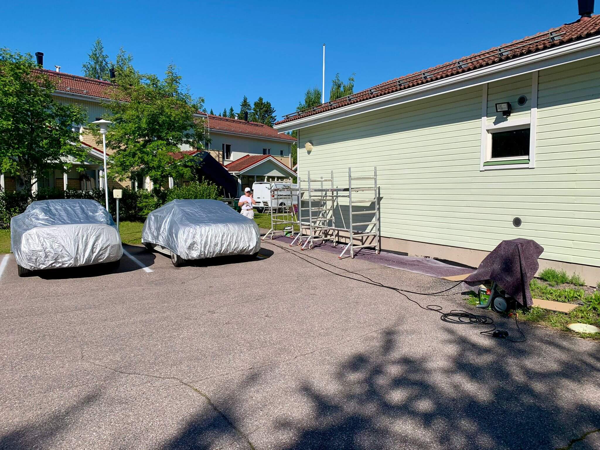 Ulkona on kaksi hopeanvärisellä suojapeitteellä peitettyä autoa, maalaustelineitä ja painepesuri. Aurinkoinen sää valaisee maisemaa.