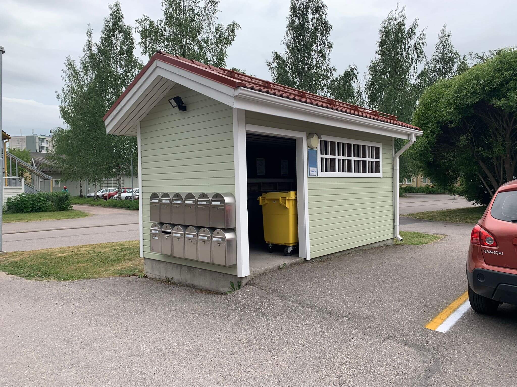 Vaaleanvihreä postilaatikkokatos punaisella katolla, jossa on metalliset postilaatikot ja keltainen roskakori, sijaitsee asfalttialueella.
