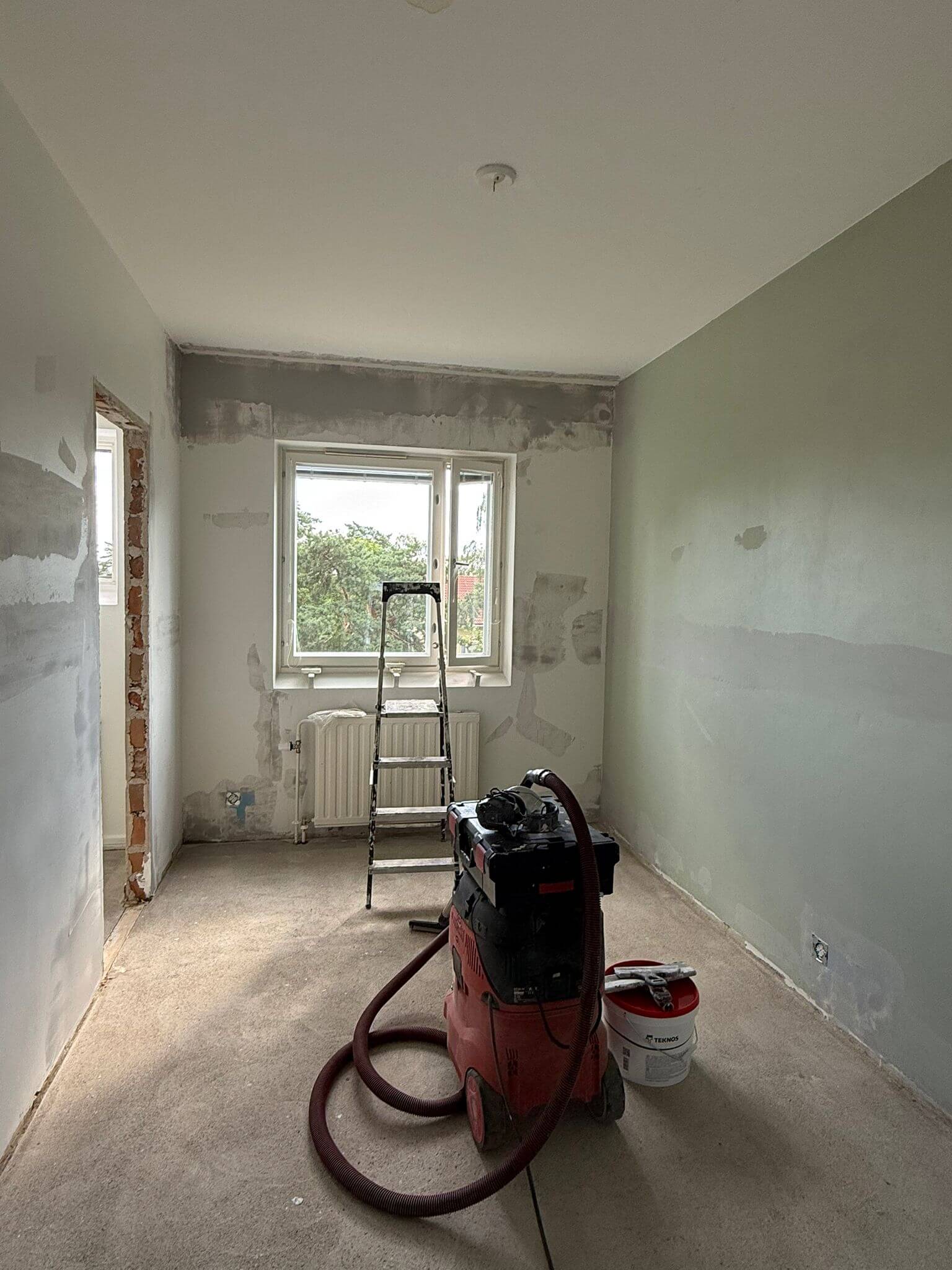Huone remontissa, jossa on tikkaat, imuri ja maalipurkki. Seinät ovat osittain maalattuja ja osittain tasoittamattomia. Valoisa huone, jossa on ikkuna.