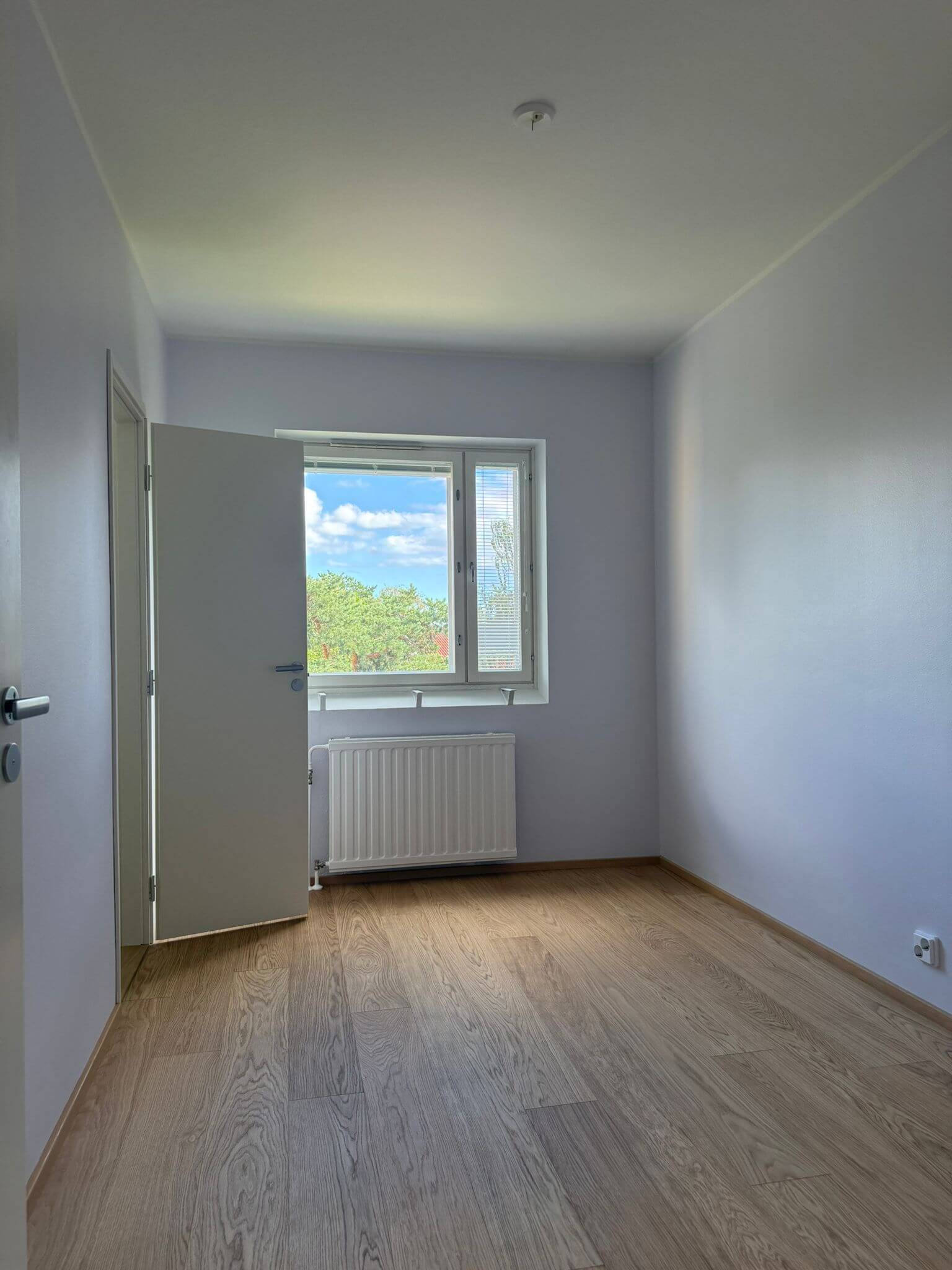 Tyhjä huone, jossa on puulattia, valkoiset seinät, avoin ovi ja ikkuna, josta näkyy taivas ja puita.