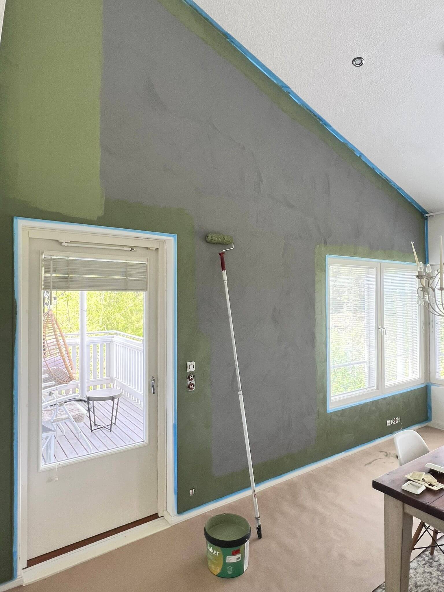 Sisätila, jossa seinä on osittain maalattu vihreäksi ja osittain harmaaksi, sinisellä maalarinteipillä rajattuna. Ikkunat ja ovi näkyvät, maalaustarvikkeet lattialla.