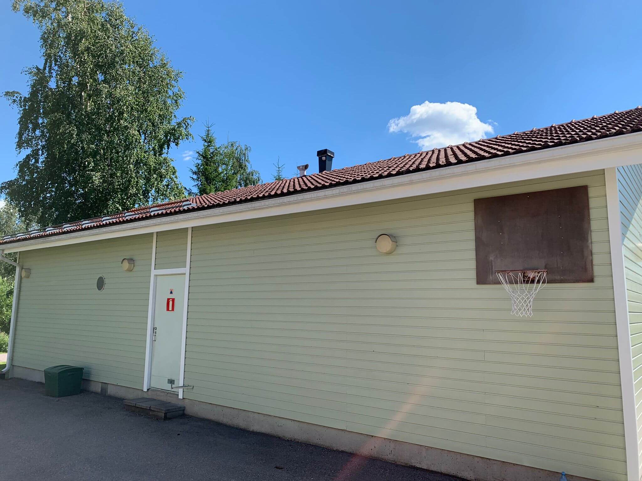 Vaaleanvihreä rakennus, jossa on ruskea katto, koripallokori ja punainen kyltti ovessa, valoisassa säässä.