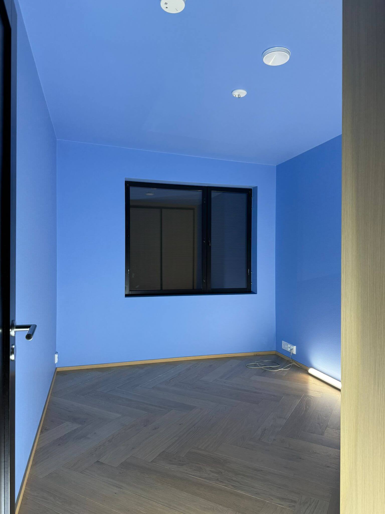 Huoneessa on siniset seinät, harmaa kalanruotokuvioinen lattia, mustakehyksinen ikkuna ja valkoiset kattovalaisimet.