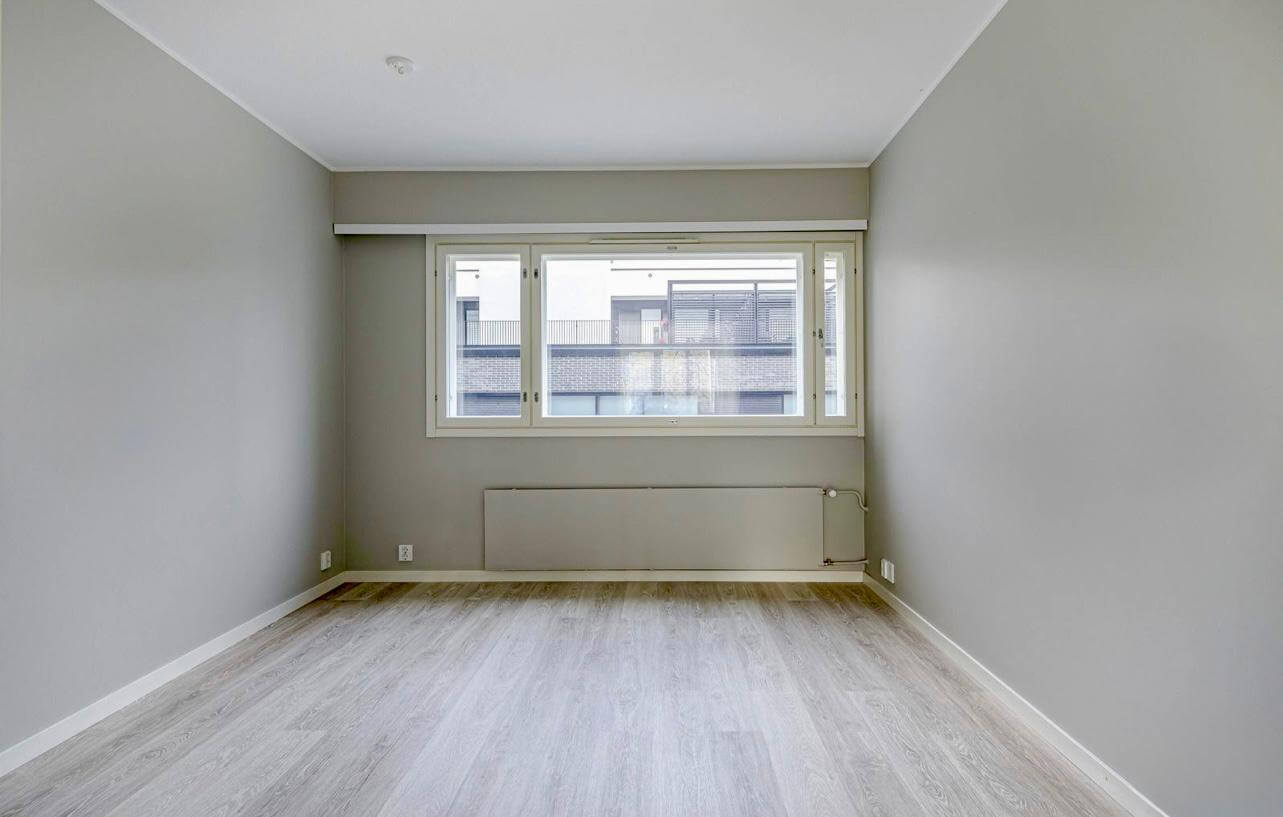 Vaaleanharmaa huone, jossa on ikkuna, laminaattilattia ja patteri. Huone on valoisa ja tyhjä.